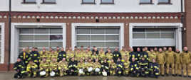 Zdjęcie grupowe dużej jednostki straży pożarnej na tle nowoczesnej remizy. Kilkudziesięciu strażaków i strażaczek ustawionych w trzech rzędach. Wszyscy są ubrani w profesjonalne mundury specjalne – przeważają stroje w kolorze piaskowym, część osób ma na sobie mundury czarne.
