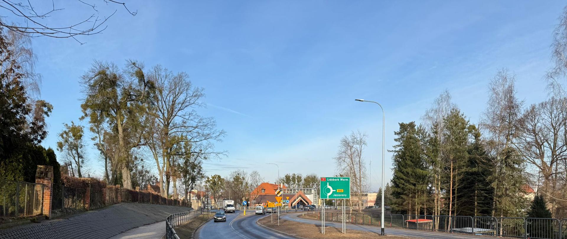 Zdjęcie przedstawia drogę krajową DK51 w Dobrym Mieście, na odcinku ul. Olsztyńskiej i ul. Cmentarnej. Widoczna jest jezdnia oddzielona od chodnika metalowymi barierkami. Na drodze porusza się kilka samochodów, w tle widać zabudowania z czerwonymi dachami oraz drzewa. Po prawej stronie znajduje się zielony drogowskaz wskazujący kierunek na Lidzbark Warmiński i przejazd przez rondo.