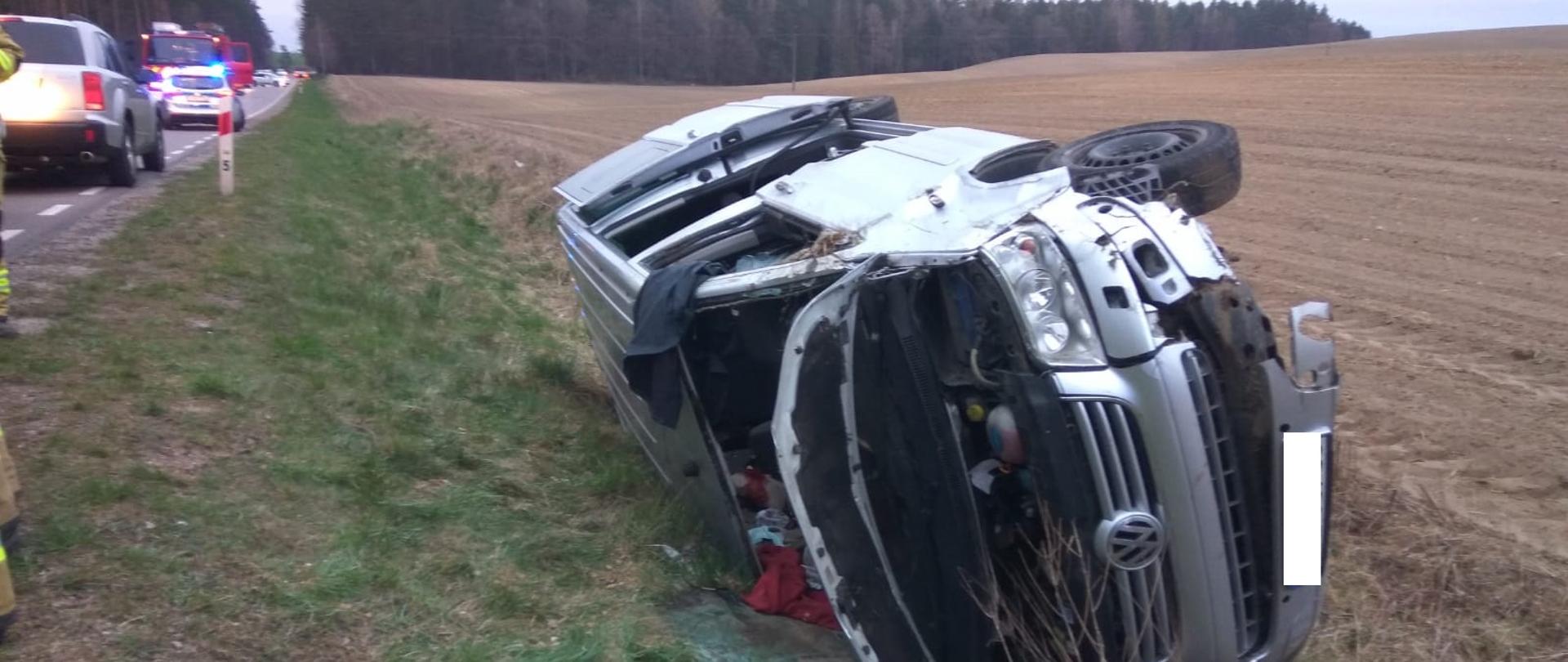 Zdjęcie przedstawia srebrne auto po wypadku. Samochód leży na poboczu drogi, na prawym boku. Ma uszkodzony zderzak, wybitą przednią szybę oraz otwarte drzwi i maskę. W tle widać drogę wiodącą przez las. Po lewej stronie zdjęcia widać drogę, na której stoi wóz strażacki.