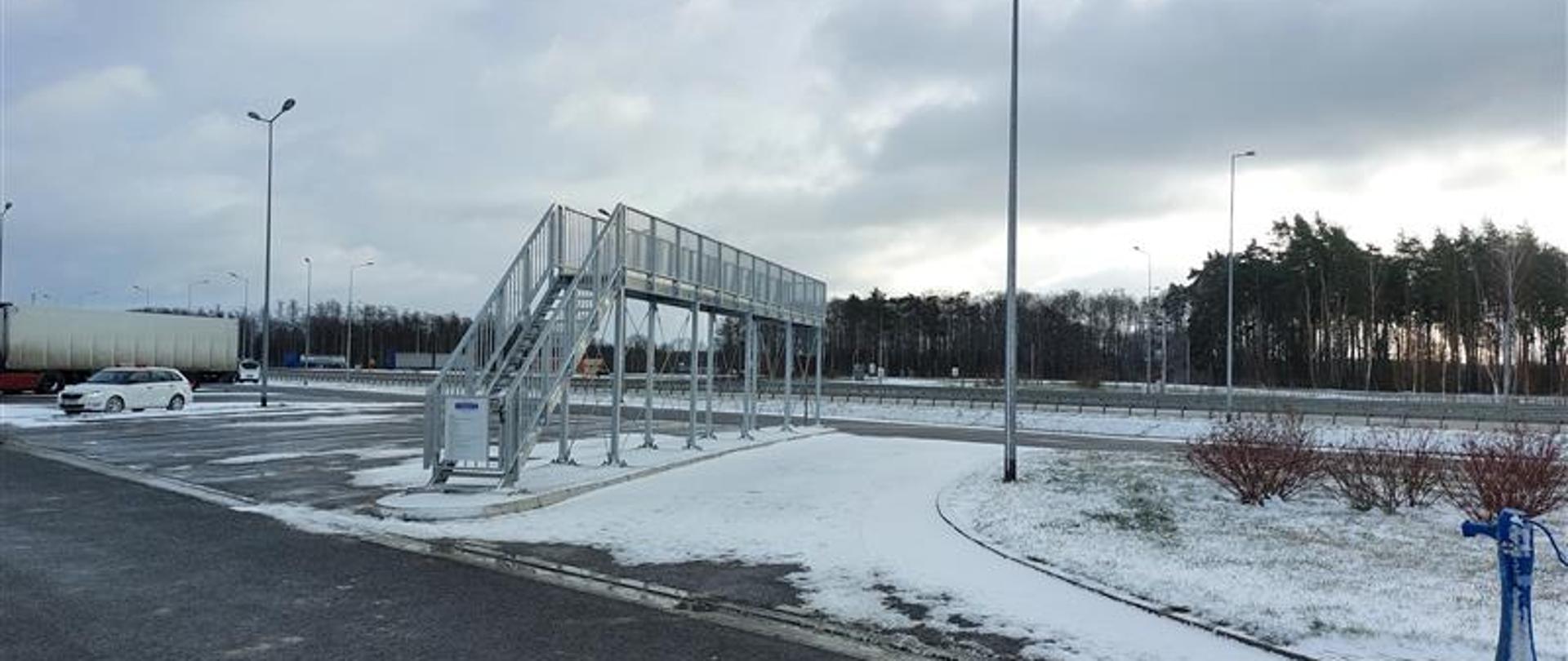 Zimowy widok na parking z metalową rampą obserwacyjną ustawioną przy krawędzi asfaltowej nawierzchni. Podłoże częściowo pokryte jest śniegiem, a w tle widać drzewa oraz zaparkowane pojazdy.