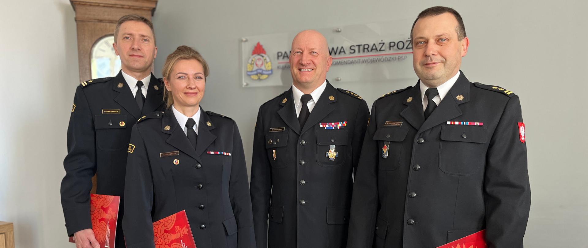 Trzech oficerów PSP w mundurach wyjściowych, w tym jedna kobieta. Stoją na tle ściany z logo Państwowej Straży Pożarnej. Troje z nich trzyma w ręku czerwoną teczkę. 