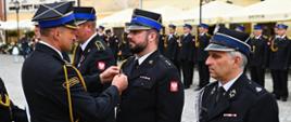 Komendant w mundurze galowym wręcza medal. W rzędzie stoją wyróżnione osoby strażacy ubrani w mundury, na klapach mają przyczepione otrzymane medale. Z tyłu widać ustawionych strażaków również w mundurach galowych.