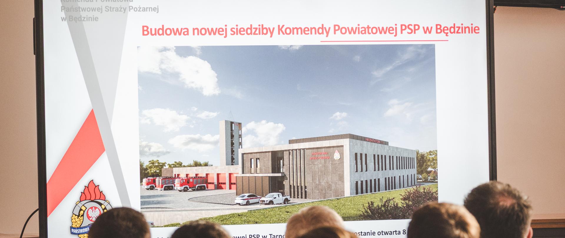 Na zdjęciu widać salę, w której grupa osób ogląda prezentację wyświetlaną na dużym ekranie. Na slajdzie znajduje się tytuł: „Budowa nowej siedziby Komendy Powiatowej PSP w Będzinie”. Centralnie umieszczono wizualizację nowoczesnego budynku straży pożarnej — obiektu z garażami na wozy bojowe, wieżą treningową oraz częścią administracyjną. Obok budynku stoją czerwone samochody pożarnicze oraz samochód osobowy.
Po lewej stronie slajdu widoczne są elementy graficzne związane z Państwową Strażą Pożarną, w tym logo PSP. W pierwszym planie widać tyły głów uczestników spotkania, którzy oglądają prezentację, co sugeruje, że wydarzenie ma charakter oficjalny lub informacyjny, np. odprawy lub konferencji dotyczącej inwestycji.
