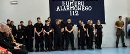 grupa dziewcząt i chłopców stoi pod ścianą na której widnieje napis Europejski Dzień Numeru Alarmowego 112 ubrani są w ubrania koszarowe służb mundurowych straży pożarnej, policji oraz wojska, po lewej stronie siedzą zaproszeni goście ubrani w ubrania służbowe pogotowia, straży pożarnej i policji
