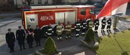 Na tle gaśniczego pojazdu widać strażaków ustawionych w szeregu do uroczystego apelu z okazji Święta Niepodległości.
Z lewej strony czterech strażaków ubranych w umundurowanie galowe jeden z nich na rękach trzyma flagę. Pozostali strażacy ubrani są w ubrania bojowe. W tle widać budynki mieszkalne i drogę.