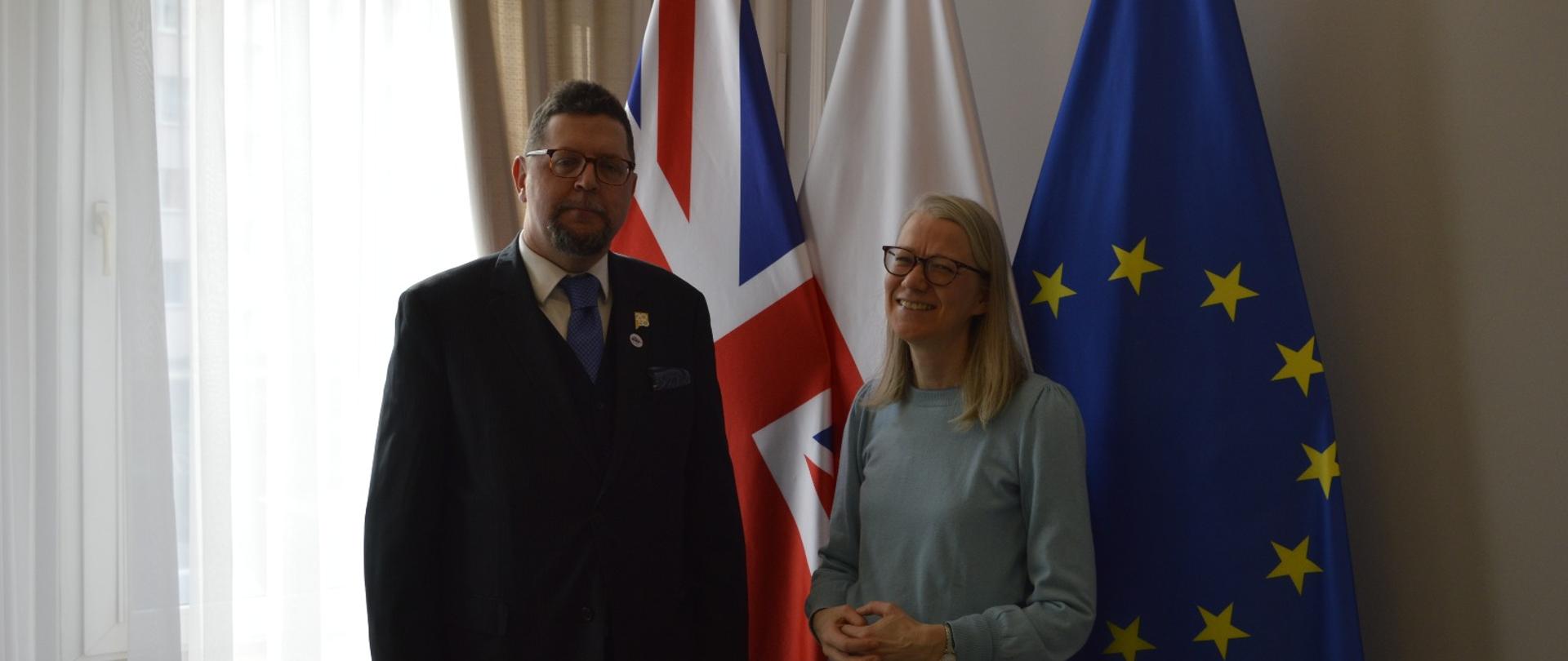 Wiceminister Szeptycki stoi obok uśmiechniętej kobiety w jasnoszarej bluzce, za nimi z lewej zasłonięte firankami okno, pod ścianą flagi Polski, UE i Wielkiej Brytanii.