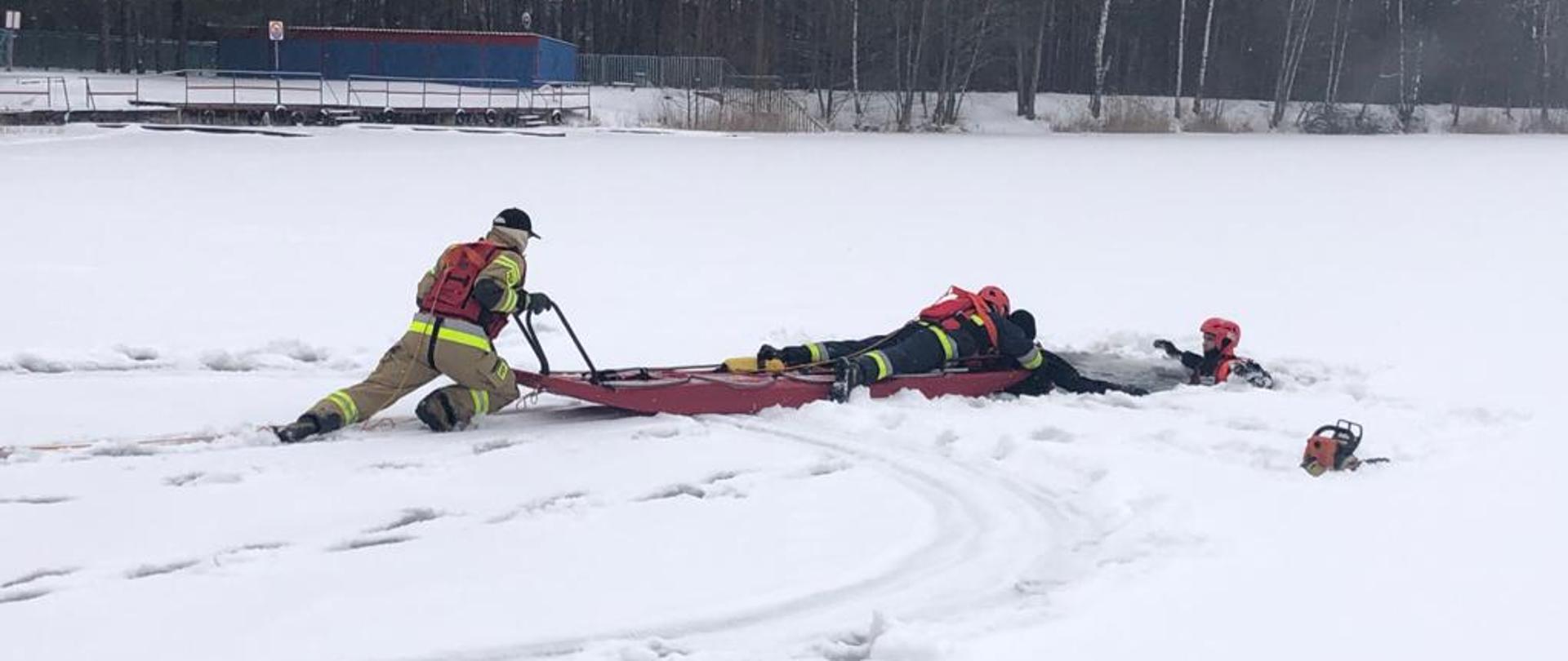 Na zdjęciu widać strażaków ćwiczących na pokrytym lodem zbiorniku wodnym. W wodzie znajduje się strażak symulujący tonącą osobę. Pozostali strażacy, wykorzystując specjalne sanie lodowe, zabezpieczeni linkami, wyciągają go z wody.
