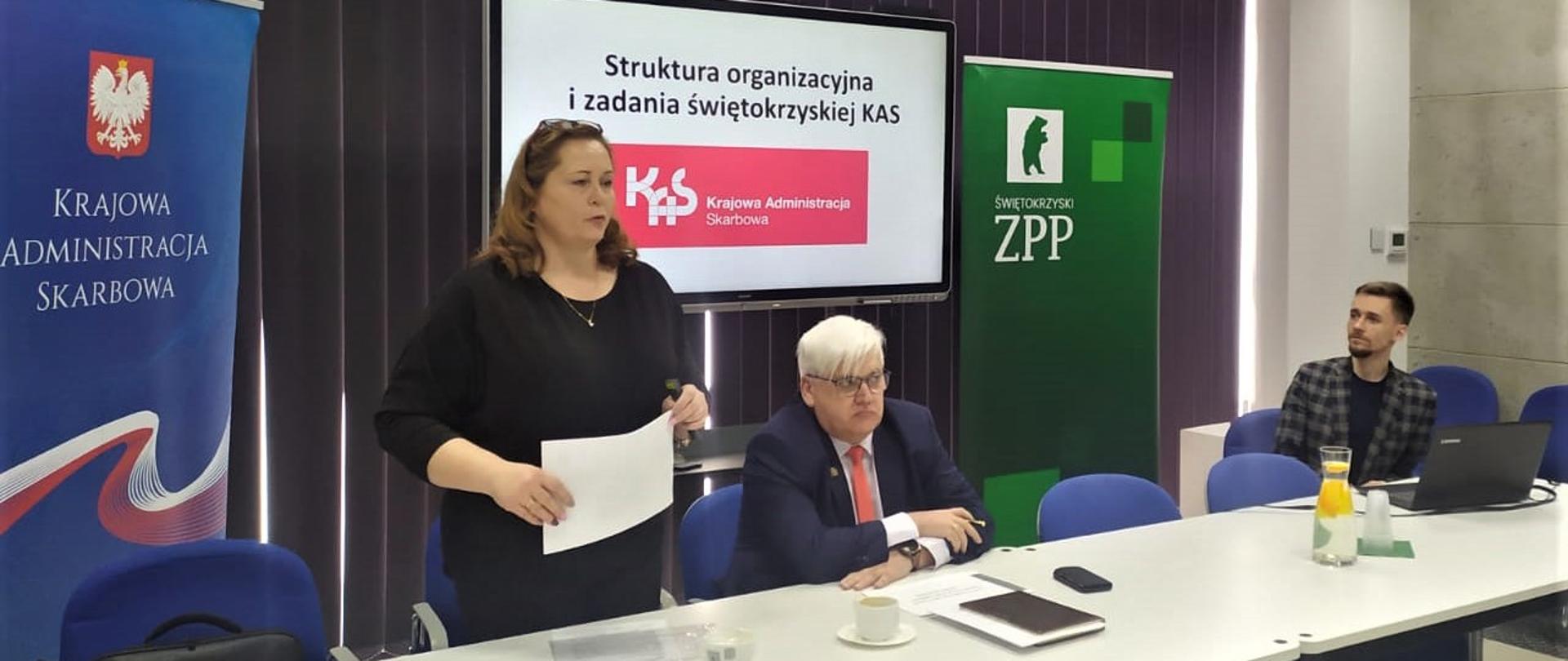 Kobieta przemawia obok przy stole konferencyjnym siedzi dwóch mężczyzn 