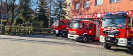Na placu zewnętrznym przed komendą stoją wystawione przed garaże samochody bojowe. Przed pierwszym z nich w szeregu stoją strażacy w mundurach bojowych.