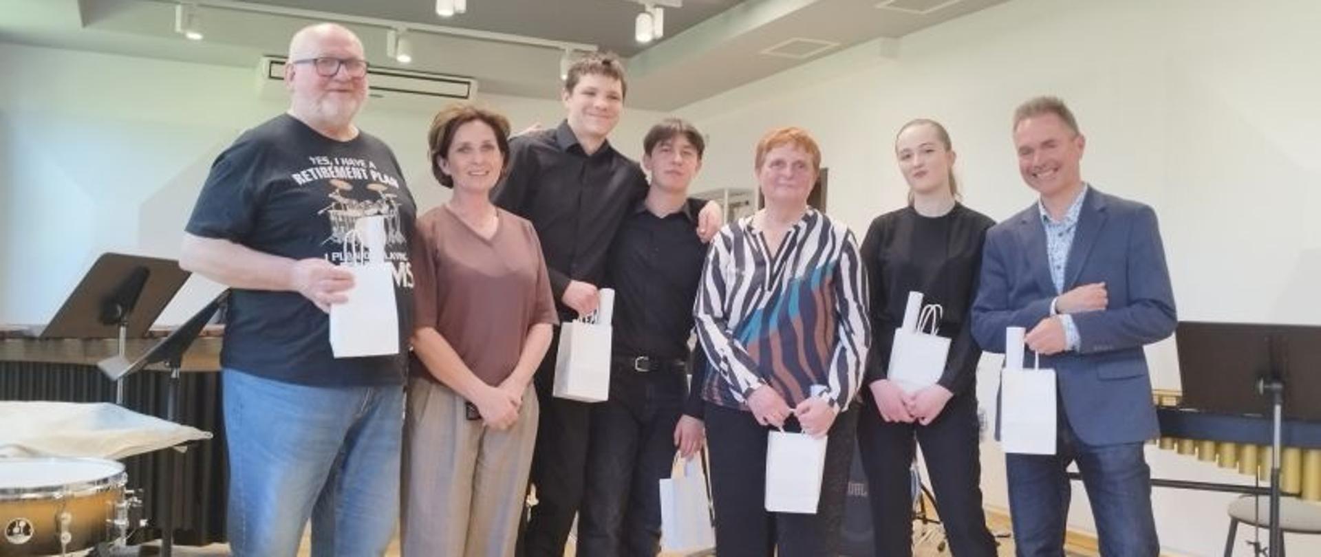 Zdjęcie kolorowe. Grupa siedmiu osób – czterech mężczyzn i trzech kobiet – stoi w rzędzie w jasnej sali z instrumentami perkusyjnymi w tle. Prawie wszyscy trzymają małe, białe torby upominkowe. Uczestnicy są ubrani w stroje wizytowe i casualowe: od czarnych koszul i marynarki po wzorzystą bluzę i koszulkę z nadrukiem. Pomieszczenie ma jasny parkiet w jodełkę i nowoczesne oświetlenie sufitowe.