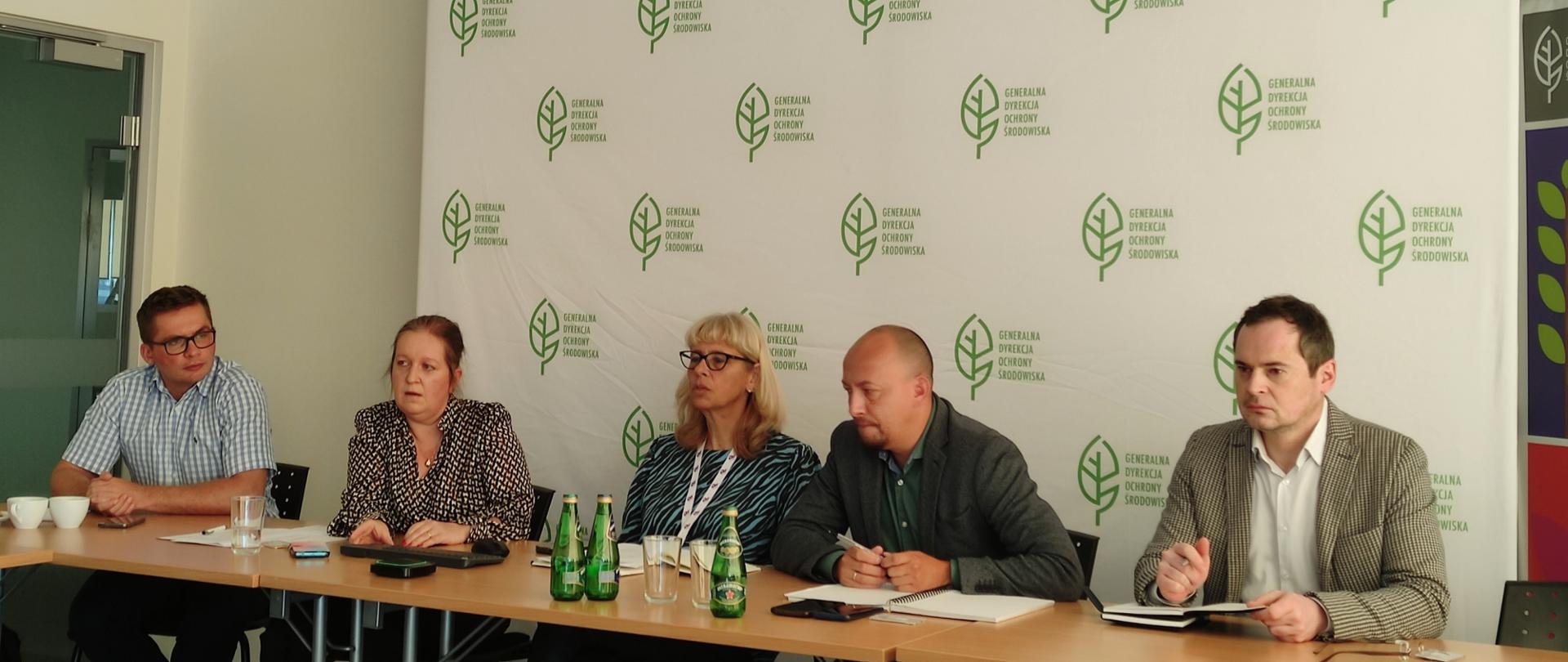 Za stołem konferencyjnym na tle białej ścianki z logotypami Generalnej Dyrekcji Ochrony Środowiska siedzą dwie kobiety i trzech mężczyzn. Niektórzy robią notatki ze spotkania. 