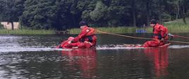 Zdjęcie wykonane jest nad jeziorem i przedstawia 3 strażaków, którzy ćwiczą techniki podejmowania osób tonących z wody. Wszyscy znajdują się w wodzie na desce i ubrani są w ubrania do pracy w wodzie oraz kamizelki ratunkowe. Widać również podpięte do strażaków linki asekuracyjne. W tle zdjęcia widać brzeg jeziora i drzewa. 
