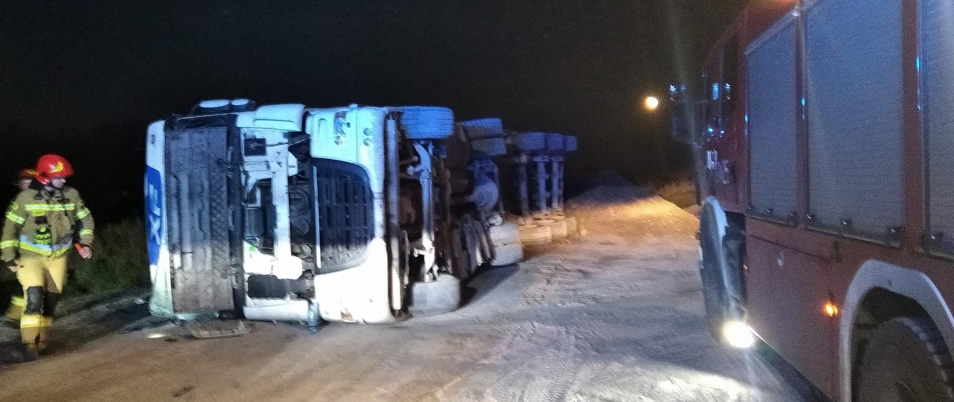 Przewrócony samochód ciężarowy na prawy bok. Przed kabiną znajduje się strażak. Z prawej strony stoi samochód pożarniczy.