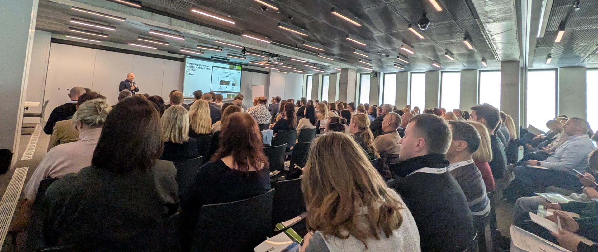 w dużej sali konferencyjnej w wielu rzędach siedzą osoby zwrócone do prowadzącego stojącego w głębi , obok wyświetlanej prezentacji