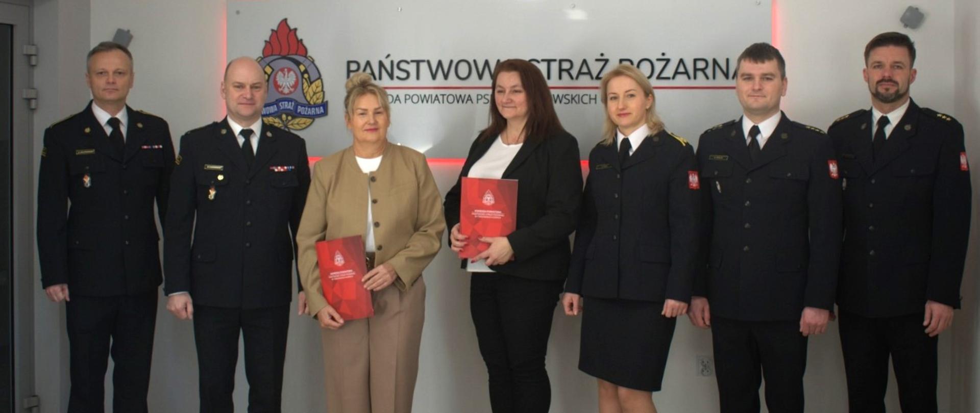 Na tle szyldu z napisem Państwowa Straż Pożarna stoi siedem osób. Czterech mężczyzn i jedna kobieta ubrani są w mundury wyjściowe. Dwie Panie w ubraniu cywilnym w ręku trzymają czerwone teczki.