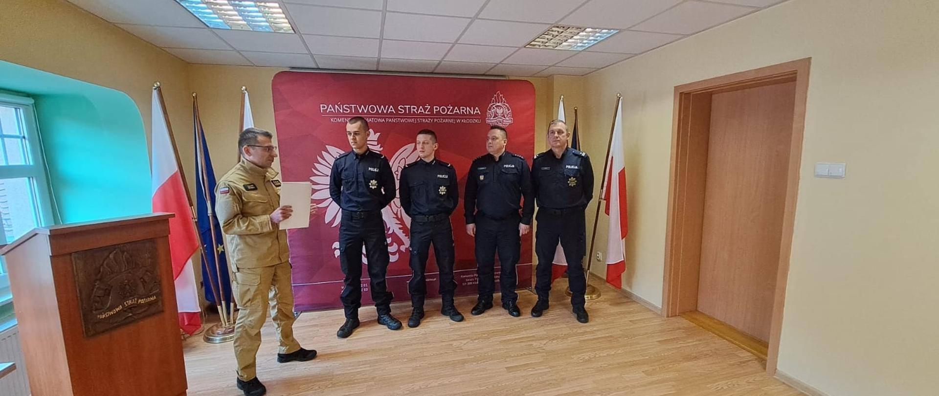 Po lewej stronie na sali konferencyjnej stoi Zastępca Komendanta Powiatowego Państwowej Straży Pożarnej w Kłodzku. Po środku stoi w rzędzie dwóch funkconariuszy policji, Komendant Powiatowy Policji w Kłodzku oraz jego Zastępca.