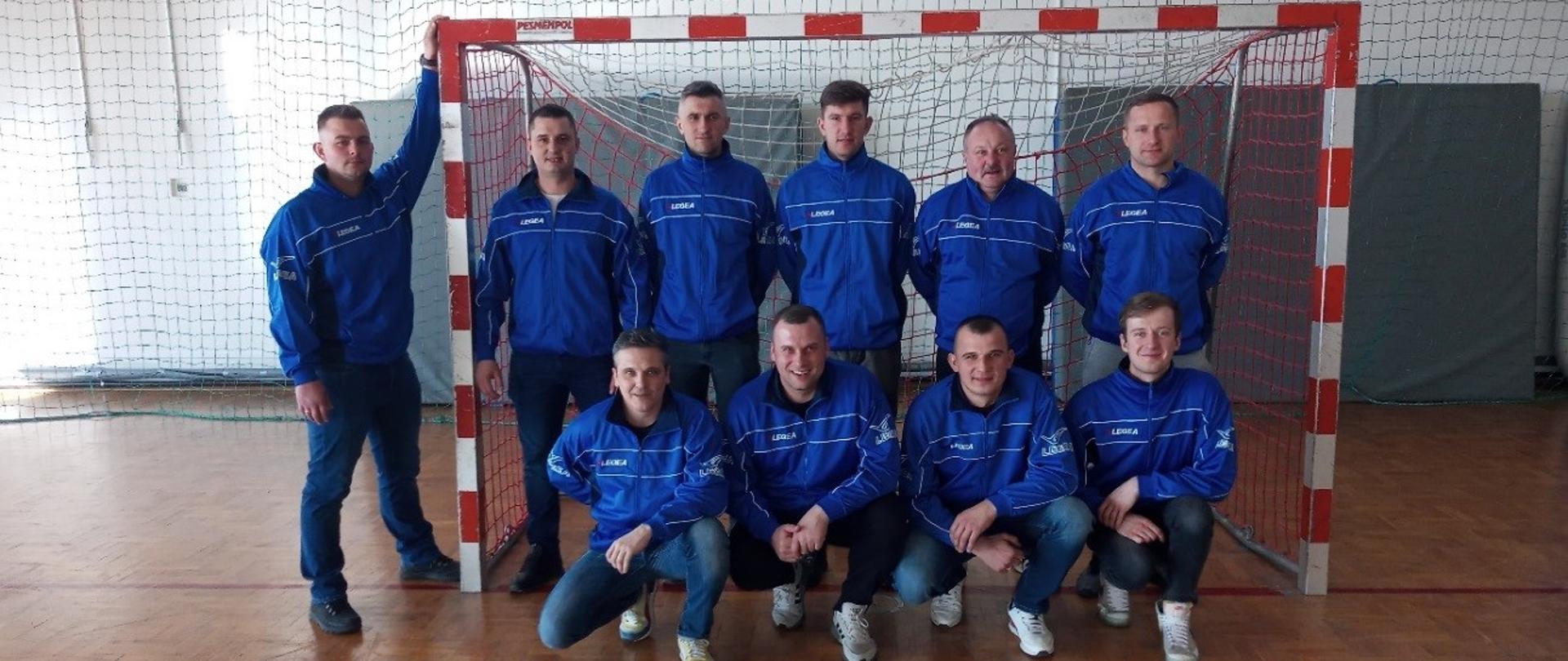 Dziesięciu sportowców ubranych w dresy koloru niebieskiego, sześciu stoi, czterech kuca na tle bramki do futsalu.