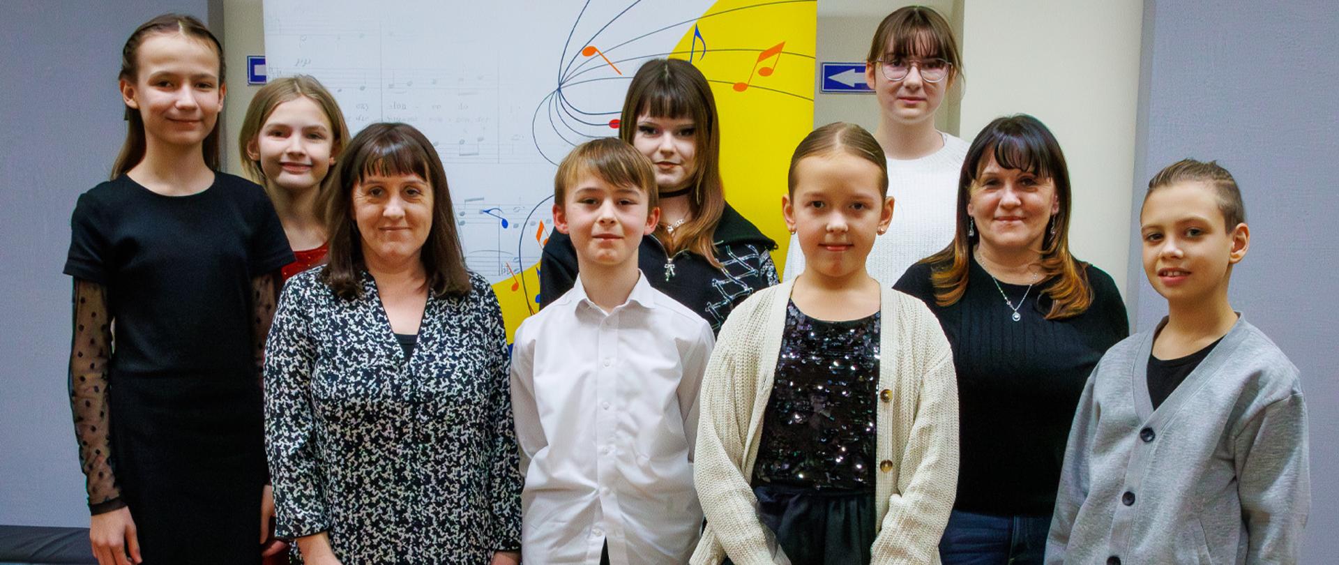 Dwie nauczycielki im kilkoro uczniów stoją na tle banera szkolnego.