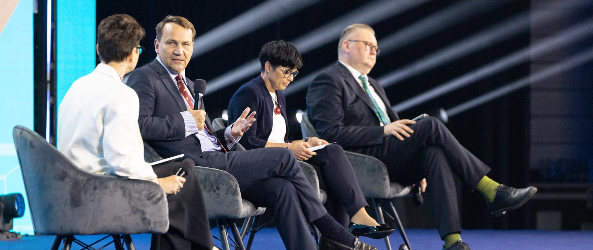 Four people take part in a panel discussion on a conference stage. Deputy Prime Minister wearing a navy suit, light blue shirt, and red tie speaks into a microphone while gesturing with his hand.