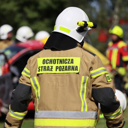Mężczyzna w stroju bojowym straży pożarnej z napisem Ochotnicza Straż Pożarna zbliża się do grupy innych strażaków przewracających rozbity samochód.