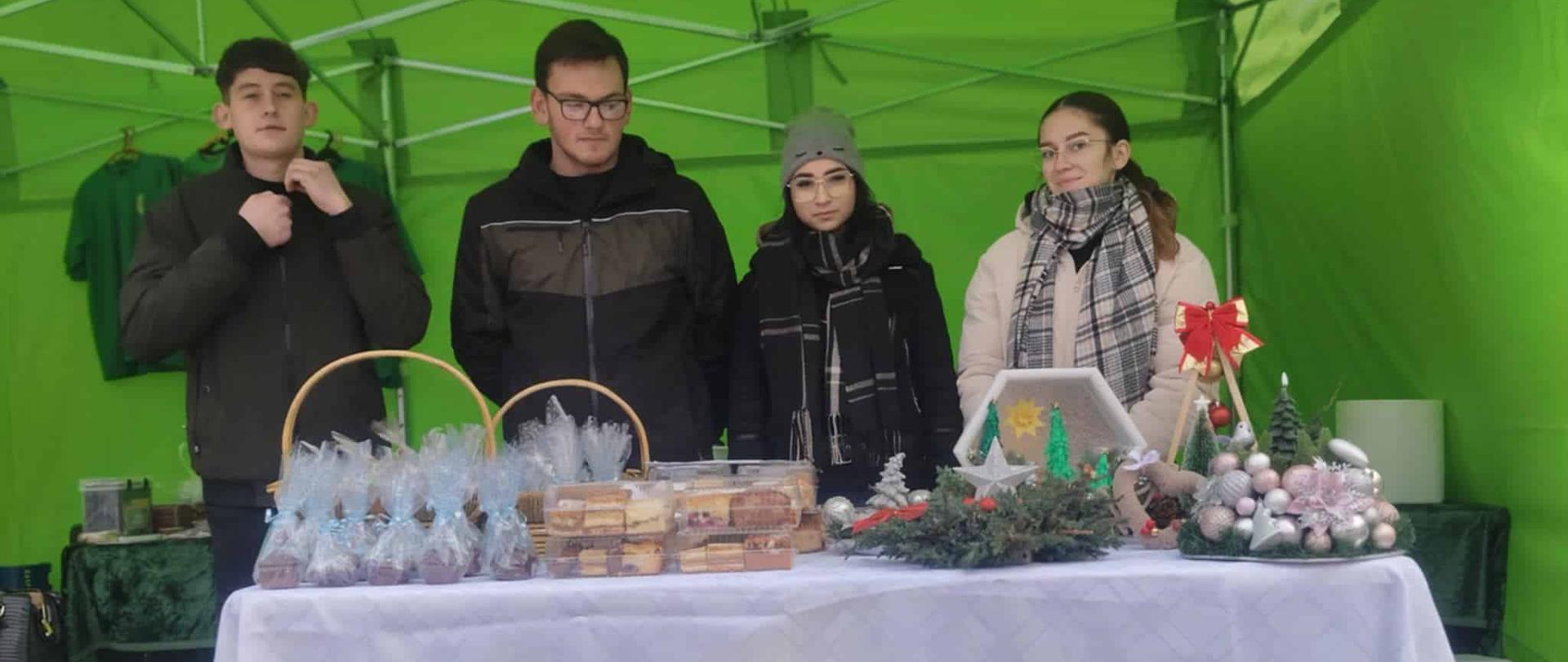 Czworo młodych ludzi stoi za ladą wewnątrz zielonego namiotu. Na białym obrusie przed nimi wyeksponowane są paczki z ciastami oraz bożonarodzeniowe dekoracje i stroiki.