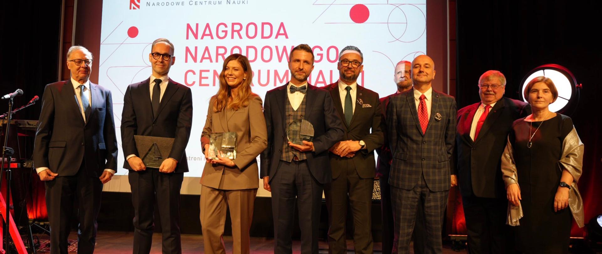 Na zaciemnionej sali przed wielkim ekranem z napisem Nagroda Narodowego Centrum Nauki stoi w rzędzie 9 osób, pośrodku grupy minister Kulasek.