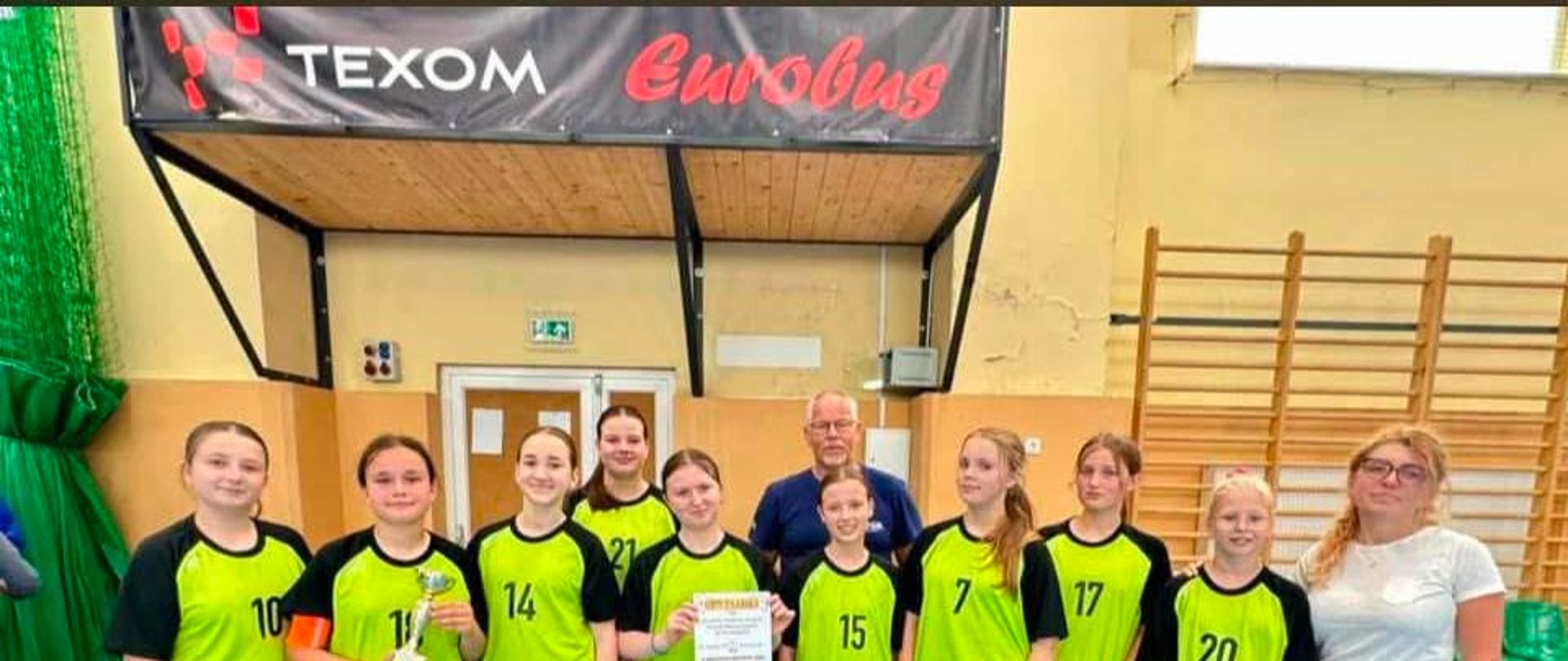 Zdjęcie grupowe dziewczęcej drużyny sportowej na hali sportowej z niebieską nawierzchnią. Osiem młodych zawodniczek stoi w rzędzie, ubranych w jaskrawozielono-czarne koszulki z numerami. Jedna z dziewcząt trzyma w dłoniach mały puchar, a druga pamiątkowy dyplom. Za nimi, pośrodku, stoi starszy mężczyzna (trener), a po prawej stronie kadru młoda kobieta w białej koszulce i dżinsach. Nad grupą wisi czarny baner z napisami „TEXOM” oraz „Eurobus”. W tle widoczne są drabinki gimnastyczne oraz drzwi wyjściowe.