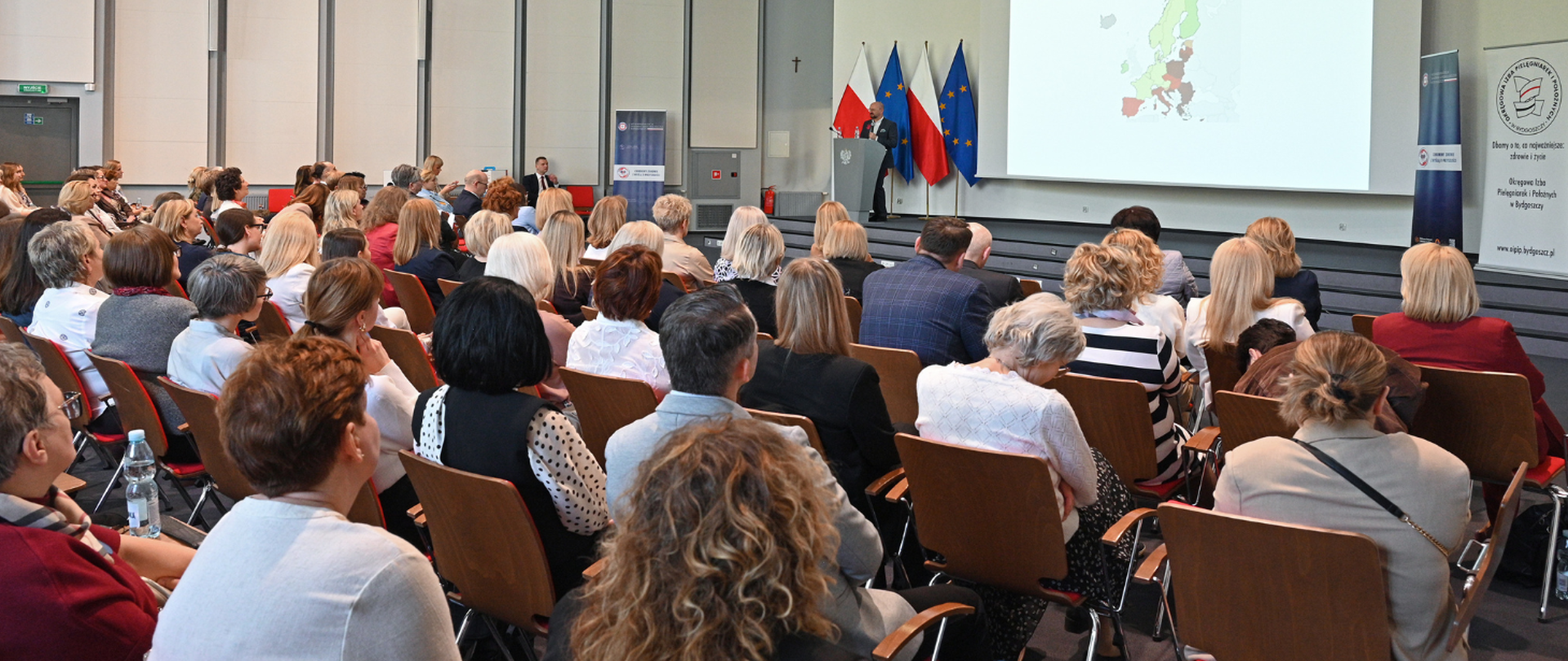 Uczestnicy konferencji siedzą w sali wykładowej i słuchają prelegenta przemawiającego przy mównicy. W tle widoczne są flagi Polski i Unii Europejskiej, ekran prezentacyjny oraz banery związane z tematyką pielęgniarstwa i higieny rąk.
